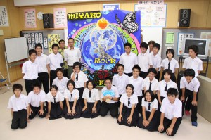 寄贈された看板幕をバックに記念撮影をする野津委員長（後列左から５人目）と生徒たち＝１９日、狩俣中学校