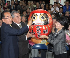 当確の知らせを受けだるまに目を入れる西銘さん（左）。右は妻の雅子さん＝16日、南風原町字山川の選対本部