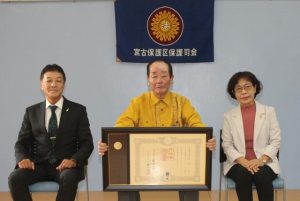 褒章の伝達を受けた奥濱さん（中央）。左は岡田所長、右は大城会長＝17日、宮古保護区保護司会サポートセンター