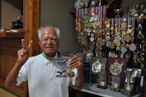 各種大会で数多くのメダルやトロフィーを獲得。今回２個のメダルが新たに加わりＶサインする亀濱さん＝18日、平良西里の自宅