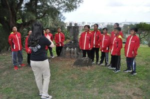 「愛と和平」の石像を挟んで合唱する子供たち＝17日、カママ嶺公園