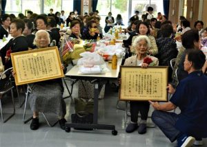 伊良部さん（前列左）、仲間さん（同右）に内閣総理大臣から賞状が伝達された＝18日、西原公民館