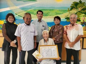 数え100歳になった本村さん（中央）に伊良皆村長から表彰状が手渡された＝15日、村コミュニティー施設