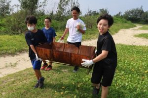 ビーチ周辺の草の中から見つかったバーベキューコンロを運ぶ児童たち＝17日、佐和田の浜