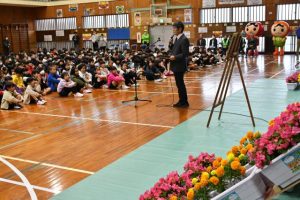 人権の花をバックにあいさつする天久校長＝９日、南小学校体育館