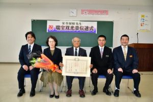 同事務所の職員が見守る中、叙勲の伝達を受けた安谷屋さん（中央）、妻の勝子さん（左２人目）、伝達した平良所長（右２人目）＝25日、県宮古合同庁舎