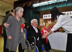 花束贈呈を受けて笑顔を見せる新里さん（中央）と恵子夫人（左）＝27日、上野公民館