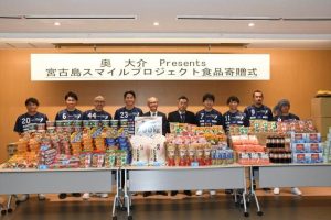 食料品を前に「スマイル」を見せる名波さん（右から４人目）、野原会長（同５人目）ら関係者の皆さん＝12日、市役所